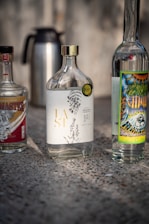 Several bottles are placed on a textured surface. The central bottle has an elegant design with botanical illustrations and gold lettering. The bottles on either side feature colorful and detailed labels. A metal thermos is seen in the background.