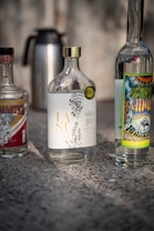 Several bottles are placed on a textured surface. The central bottle has an elegant design with botanical illustrations and gold lettering. The bottles on either side feature colorful and detailed labels. A metal thermos is seen in the background.