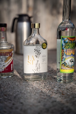 Several bottles are placed on a textured surface. The central bottle has an elegant design with botanical illustrations and gold lettering. The bottles on either side feature colorful and detailed labels. A metal thermos is seen in the background.