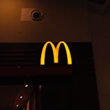 A prominently lit yellow 'M' logo on a dark background, characteristic of a well-known fast-food chain.