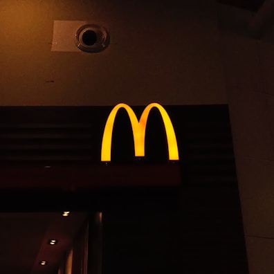 A prominently lit yellow 'M' logo on a dark background, characteristic of a well-known fast-food chain.