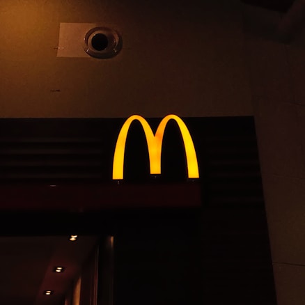 A prominently lit yellow 'M' logo on a dark background, characteristic of a well-known fast-food chain.