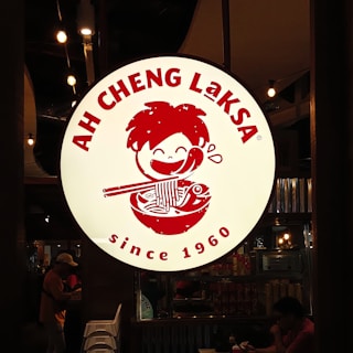 A large, circular sign featuring a playful illustration of a person eating noodles. The text 'AH CHENG LAKSA' is prominently displayed around the top edge, and 'since 1960' is written on the bottom. The sign is brightly illuminated against a dimly lit interior background with people and chairs visible.