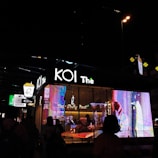 A nighttime street scene featuring a KOI Thé bubble tea shop, illuminated by bright, colorful digital displays and signage. Silhouettes of people can be seen in front of the shop, creating a bustling urban atmosphere.