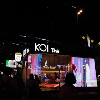 A nighttime street scene featuring a KOI Thé bubble tea shop, illuminated by bright, colorful digital displays and signage. Silhouettes of people can be seen in front of the shop, creating a bustling urban atmosphere.
