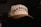 A cream-colored baseball cap with black embroidered text reading 'DADGANG' and a smaller line saying 'Designed by Dads'. The cap is resting on a dark surface with a dimly lit background. The text is bold and prominent, giving it a stylish and personalized look.