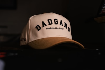 A cream-colored baseball cap with black embroidered text reading 'DADGANG' and a smaller line saying 'Designed by Dads'. The cap is resting on a dark surface with a dimly lit background. The text is bold and prominent, giving it a stylish and personalized look.