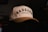 A cream-colored baseball cap with black embroidered text reading 'DADGANG' and a smaller line saying 'Designed by Dads'. The cap is resting on a dark surface with a dimly lit background. The text is bold and prominent, giving it a stylish and personalized look.