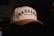 A cream-colored baseball cap with black embroidered text reading 'DADGANG' and a smaller line saying 'Designed by Dads'. The cap is resting on a dark surface with a dimly lit background. The text is bold and prominent, giving it a stylish and personalized look.