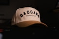 A cream-colored baseball cap with black embroidered text reading 'DADGANG' and a smaller line saying 'Designed by Dads'. The cap is resting on a dark surface with a dimly lit background. The text is bold and prominent, giving it a stylish and personalized look.