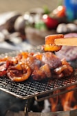 Pieces of grilled meat are sizzling on a wire-mesh grill over an open flame. A piece of meat, marinated and browned, is held by chopsticks in the foreground, with a blurred vegetable background suggesting a barbecue setting.