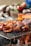 Pieces of grilled meat are sizzling on a wire-mesh grill over an open flame. A piece of meat, marinated and browned, is held by chopsticks in the foreground, with a blurred vegetable background suggesting a barbecue setting.