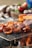 Pieces of grilled meat are sizzling on a wire-mesh grill over an open flame. A piece of meat, marinated and browned, is held by chopsticks in the foreground, with a blurred vegetable background suggesting a barbecue setting.