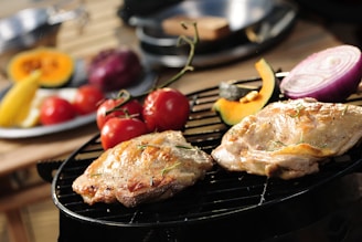 Two pieces of seasoned chicken are grilling on a barbecue, alongside fresh cherry tomatoes, a slice of red onion, and a wedge of squash. In the background, there is a blurred plate with various vegetables, including a red onion and a banana.