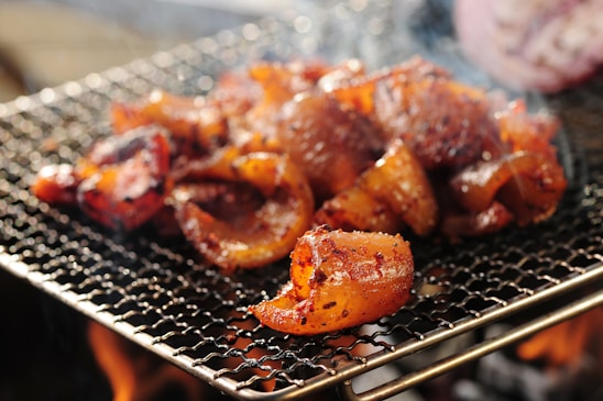 Pieces of marinated meat are cooking on a grill, with visible char marks and a smoky background. The meat is sizzling, indicating it's on an active barbecue with flames visible beneath the grill.