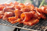 Sizzling pieces of marinated meat are cooking on a barbecue grill. The meat is well-seasoned with a rich, red marinade, and it's placed on a wire mesh grill. The background is slightly blurred, emphasizing the focus on the meat with hints of smoke rising