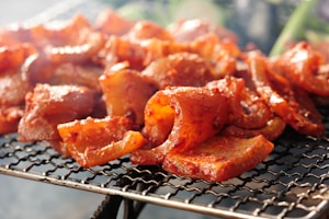 Sizzling pieces of marinated meat are cooking on a barbecue grill. The meat is well-seasoned with a rich, red marinade, and it's placed on a wire mesh grill. The background is slightly blurred, emphasizing the focus on the meat with hints of smoke rising