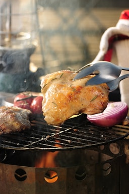 A piece of grilled chicken is being lifted with a fork over a barbecue grill. The grill also holds other food items, including a red onion half and what appears to be a tomato. Flames and smoke rise from the grill, suggesting a cooking process.