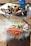 A barbecue grill is cooking seasoned meat alongside sliced vegetables which include zucchini, eggplant, asparagus, and onion. In the background, there's a table set with bowls containing fresh vegetables like tomatoes, bell peppers, and mushrooms, along with a gas canister and a spice container.