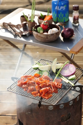 A barbecue grill is cooking seasoned meat alongside sliced vegetables which include zucchini, eggplant, asparagus, and onion. In the background, there's a table set with bowls containing fresh vegetables like tomatoes, bell peppers, and mushrooms, along with a gas canister and a spice container.