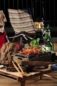 A wooden table holds a small grill with cooked, seasoned food, likely chicken. Surrounding the grill are fresh vegetables and a couple of bottles of beverage. In the background, a colorful, striped chair, a red chair with a towel draped over it, and an old-fashioned lantern are visible.