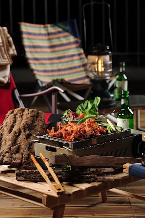 A wooden table holds a small grill with cooked, seasoned food, likely chicken. Surrounding the grill are fresh vegetables and a couple of bottles of beverage. In the background, a colorful, striped chair, a red chair with a towel draped over it, and an old-fashioned lantern are visible.