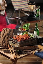 Outdoor picnic setting featuring a grill with sizzling marinated meat and vegetables. Wooden tables support the grill and various items, including tongs and wooden logs. A salad bowl with fresh leafy greens and assorted colorful vegetables sits nearby. A bottle of green glass, resembling a soju bottle, and a lantern add to the camping atmosphere. Foldable chairs with patterned and solid fabric backrests accompany the setup.