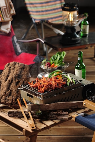 Outdoor picnic setting featuring a grill with sizzling marinated meat and vegetables. Wooden tables support the grill and various items, including tongs and wooden logs. A salad bowl with fresh leafy greens and assorted colorful vegetables sits nearby. A bottle of green glass, resembling a soju bottle, and a lantern add to the camping atmosphere. Foldable chairs with patterned and solid fabric backrests accompany the setup.