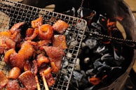 Grilled meat pieces arranged on a wire mesh over glowing charcoal. The meat appears to be well-seasoned and cooked, with a slightly glossy and caramelized finish. The charcoal beneath is dark and partially lit, with visible orange flames.