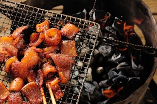 Grilled meat pieces arranged on a wire mesh over glowing charcoal. The meat appears to be well-seasoned and cooked, with a slightly glossy and caramelized finish. The charcoal beneath is dark and partially lit, with visible orange flames.