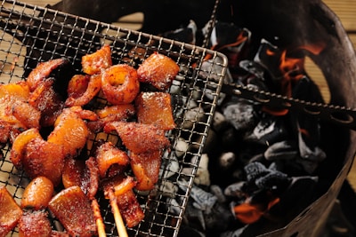 Grilled meat pieces arranged on a wire mesh over glowing charcoal. The meat appears to be well-seasoned and cooked, with a slightly glossy and caramelized finish. The charcoal beneath is dark and partially lit, with visible orange flames.