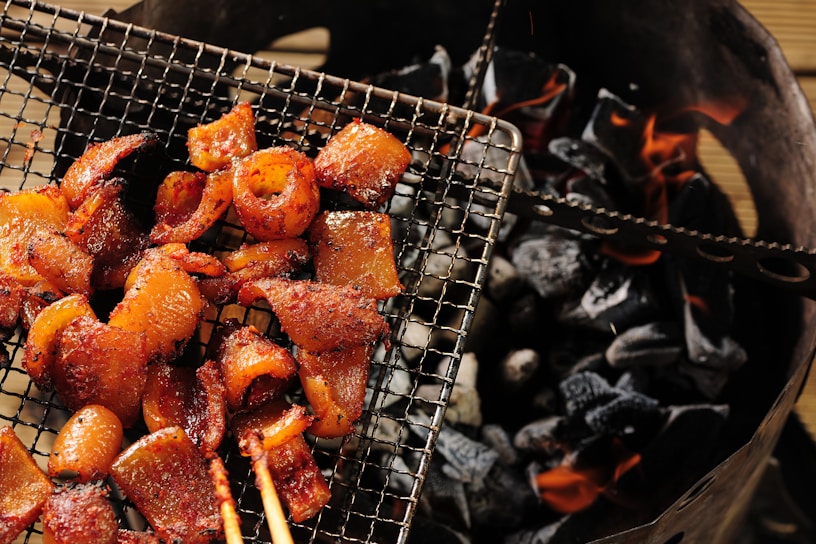 Grilled meat pieces arranged on a wire mesh over glowing charcoal. The meat appears to be well-seasoned and cooked, with a slightly glossy and caramelized finish. The charcoal beneath is dark and partially lit, with visible orange flames.