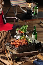 A tabletop grill laden with skewered and grilled meat, garnished with green peppers. A fresh salad bowl with leafy greens and sliced vegetables sits nearby. A bottle of green glass and a lantern are placed on a dark surface in the background. Wooden logs and chopsticks are arranged around the grilling area, and a red camping chair is partially visible.