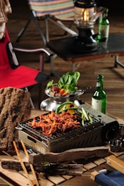 A tabletop grill laden with skewered and grilled meat, garnished with green peppers. A fresh salad bowl with leafy greens and sliced vegetables sits nearby. A bottle of green glass and a lantern are placed on a dark surface in the background. Wooden logs and chopsticks are arranged around the grilling area, and a red camping chair is partially visible.