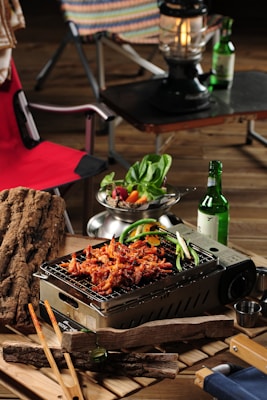 A tabletop grill laden with skewered and grilled meat, garnished with green peppers. A fresh salad bowl with leafy greens and sliced vegetables sits nearby. A bottle of green glass and a lantern are placed on a dark surface in the background. Wooden logs and chopsticks are arranged around the grilling area, and a red camping chair is partially visible.