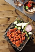 A grill placed on a wooden deck laden with assorted vegetables and grilled meat. Sliced pieces of marinated meat are cooked alongside vegetables including asparagus, brussels sprouts, eggplant, and a ring of purple onion. Nearby, a table holds a wooden bowl filled with fresh tomatoes on the vine, mushrooms, and more brussels sprouts, while wooden utensils are scattered next to it.