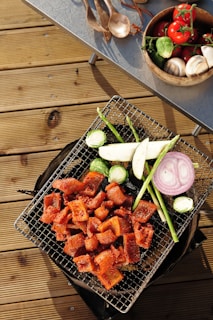 A grill placed on a wooden deck laden with assorted vegetables and grilled meat. Sliced pieces of marinated meat are cooked alongside vegetables including asparagus, brussels sprouts, eggplant, and a ring of purple onion. Nearby, a table holds a wooden bowl filled with fresh tomatoes on the vine, mushrooms, and more brussels sprouts, while wooden utensils are scattered next to it.