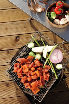 A grill placed on a wooden deck laden with assorted vegetables and grilled meat. Sliced pieces of marinated meat are cooked alongside vegetables including asparagus, brussels sprouts, eggplant, and a ring of purple onion. Nearby, a table holds a wooden bowl filled with fresh tomatoes on the vine, mushrooms, and more brussels sprouts, while wooden utensils are scattered next to it.