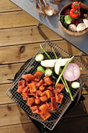 A grill placed on a wooden deck laden with assorted vegetables and grilled meat. Sliced pieces of marinated meat are cooked alongside vegetables including asparagus, brussels sprouts, eggplant, and a ring of purple onion. Nearby, a table holds a wooden bowl filled with fresh tomatoes on the vine, mushrooms, and more brussels sprouts, while wooden utensils are scattered next to it.