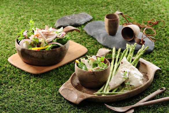 A rustic outdoor setting displays two wooden plates on a grassy surface, each holding a different dish. On the left, there is a fresh salad with a variety of colorful vegetables and shredded chicken. On the right, the plate features a piece of white fish topped with herbs, accompanied by asparagus spears and a smaller side salad. In the background, there are wooden cups on slate stones.