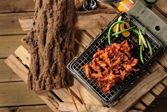 Grilled marinated meat on a wire rack, garnished with green onions and slices of orange, set on a rustic wooden surface. A textured piece of wood is placed to the side, adding to the natural, earthy theme.
