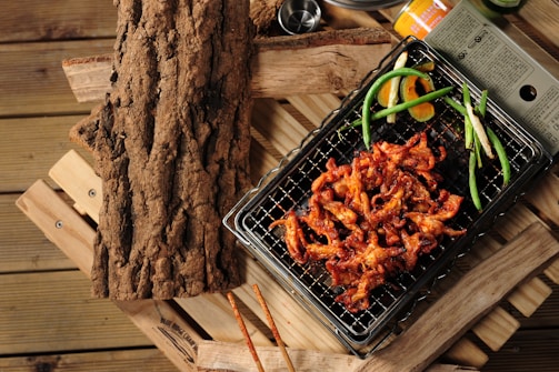 Grilled marinated meat on a wire rack, garnished with green onions and slices of orange, set on a rustic wooden surface. A textured piece of wood is placed to the side, adding to the natural, earthy theme.