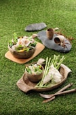 Two rustic bowls filled with fresh salads featuring sliced vegetables and meats are placed on a grassy surface. Beside the bowls, there is cutlery and stone plates with earthenware cups, creating a natural and organic dining setup. Asparagus spears and a piece of white fish with pepper seasoning are prominently displayed.