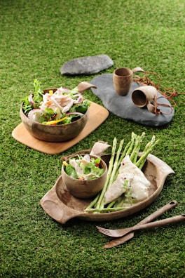 Two rustic bowls filled with fresh salads featuring sliced vegetables and meats are placed on a grassy surface. Beside the bowls, there is cutlery and stone plates with earthenware cups, creating a natural and organic dining setup. Asparagus spears and a piece of white fish with pepper seasoning are prominently displayed.