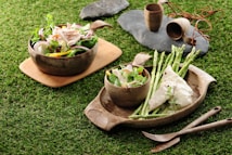 A rustic table setting features a wooden platter with fresh salad and chicken alongside asparagus. Rough stone surfaces hold clay mugs and natural rope for an earthy feel. The scene is set on vibrant grass, emphasizing an outdoor, picnic-like atmosphere.