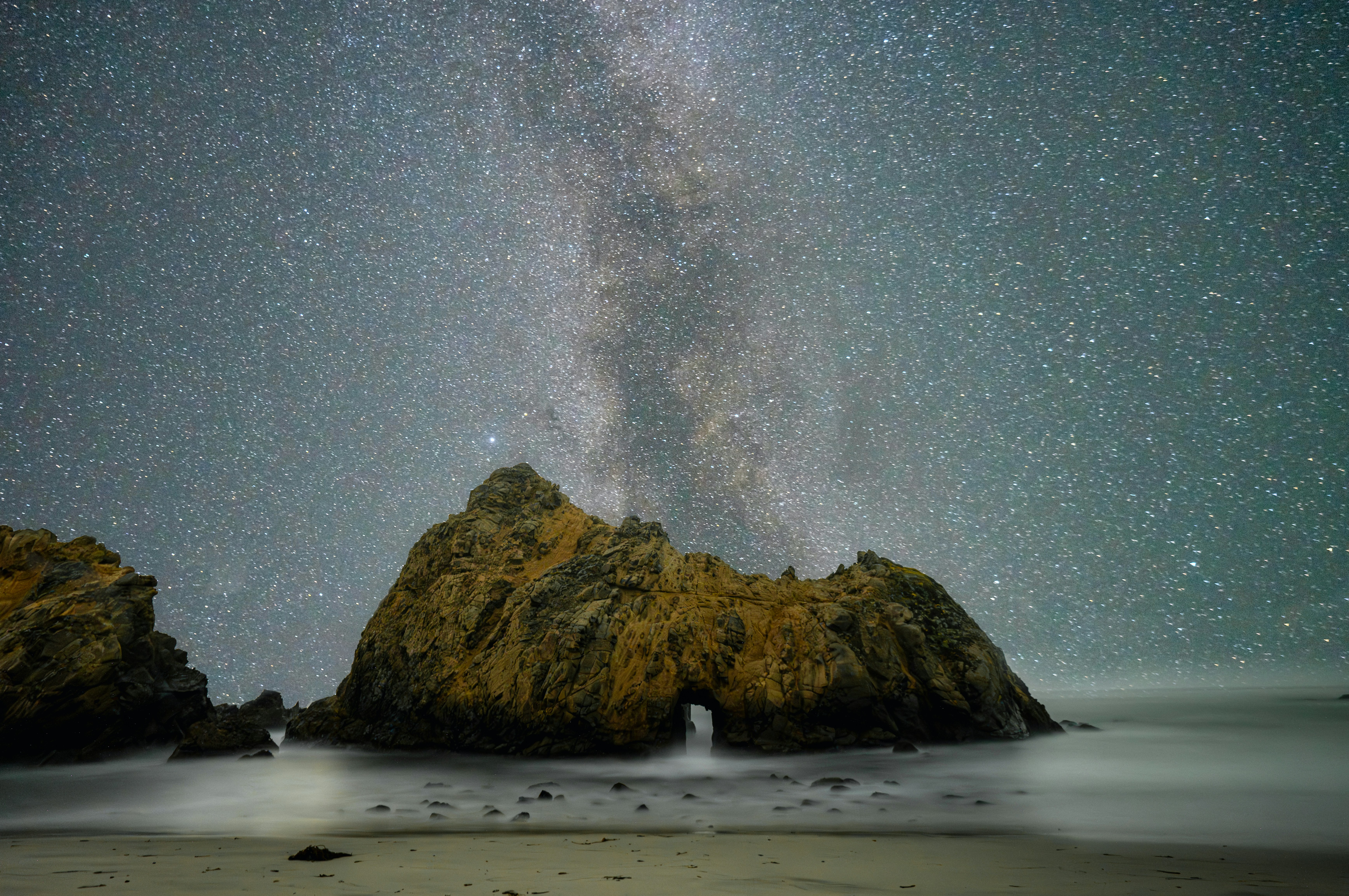 a large rock sitting on top of a beach under a sky filled with stars, 