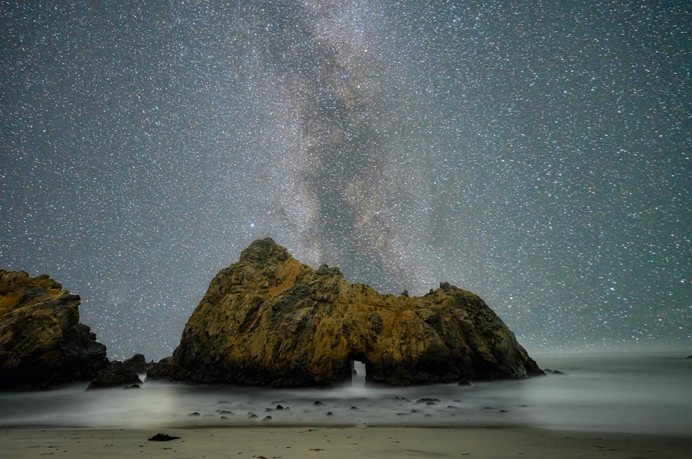 a large rock sitting on top of a beach under a sky filled with stars