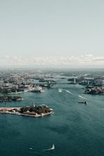 An aerial view of a bustling harbor with a prominent bridge and an iconic opera house. The water is dotted with various boats, and the skyline of the city extends into the distance under a pale blue sky.