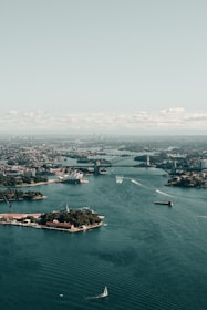 An aerial view of a bustling harbor with a prominent bridge and an iconic opera house. The water is dotted with various boats, and the skyline of the city extends into the distance under a pale blue sky.