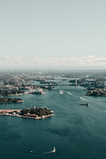 An aerial view of a bustling harbor with a prominent bridge and an iconic opera house. The water is dotted with various boats, and the skyline of the city extends into the distance under a pale blue sky.